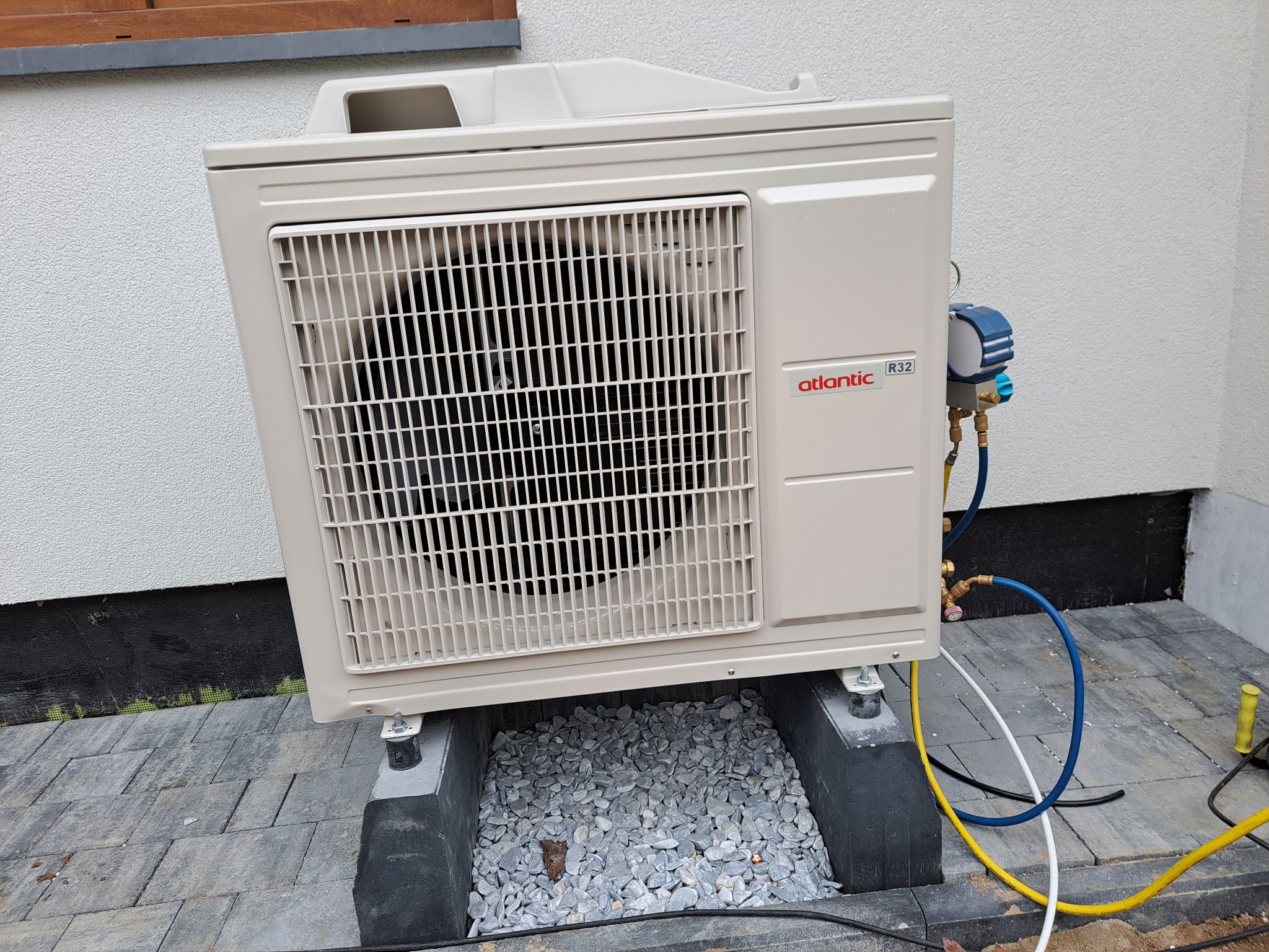 Zewnętrzna jednostka klimatyzacji marki Atlantic R32, zamontowana na betonowych podstawach z wypełnieniem z szarego żwiru, podłączona do manometru z przewodami w kolorach żółtym i niebieskim,...