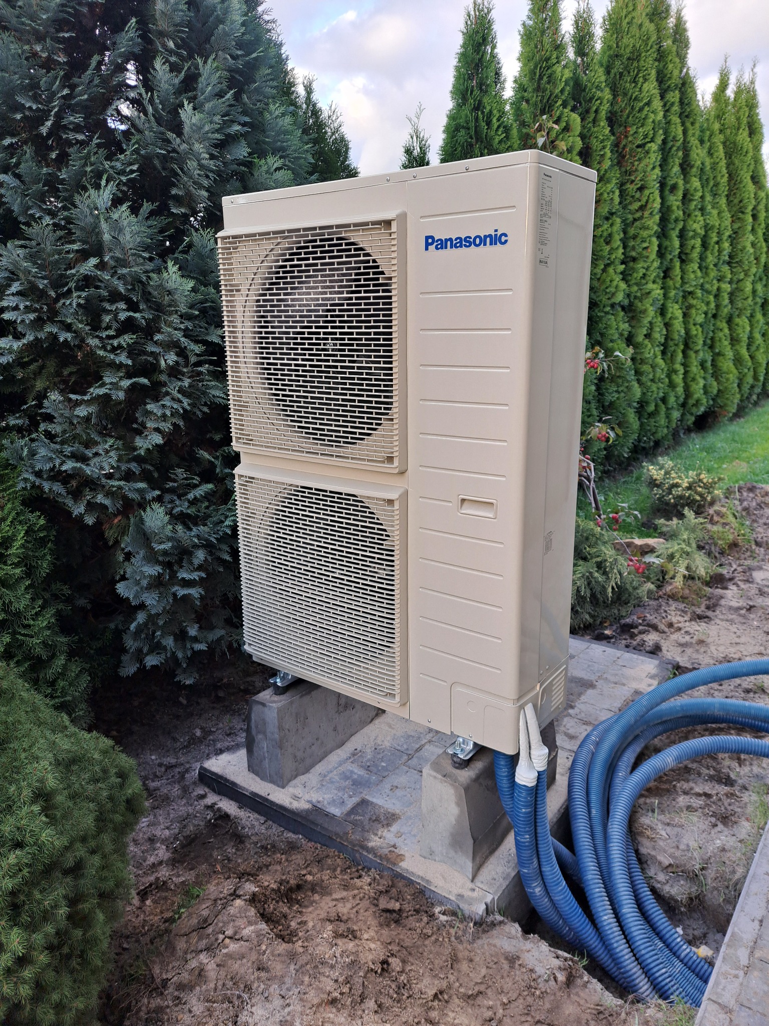 Zewnętrzna jednostka klimatyzacji Panasonic zamontowana na betonowych podstawach w ogrodzie, z widocznymi niebieskimi przewodami instalacyjnymi.