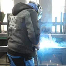 Spawacz w masce ochronnej łączy elementy metalowej konstrukcji, widoczne iskry i dym spawalniczy, na plecach bluzy logo DC.