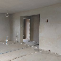 Usługi tynkarskie Adam Nowak - Świeżo otynkowane wnętrze z widocznymi instalacjami podłogowymi i otworem w ścianie, gotowe do dalszych prac wykończeniowych. Szpachla na ścianach i suficie.