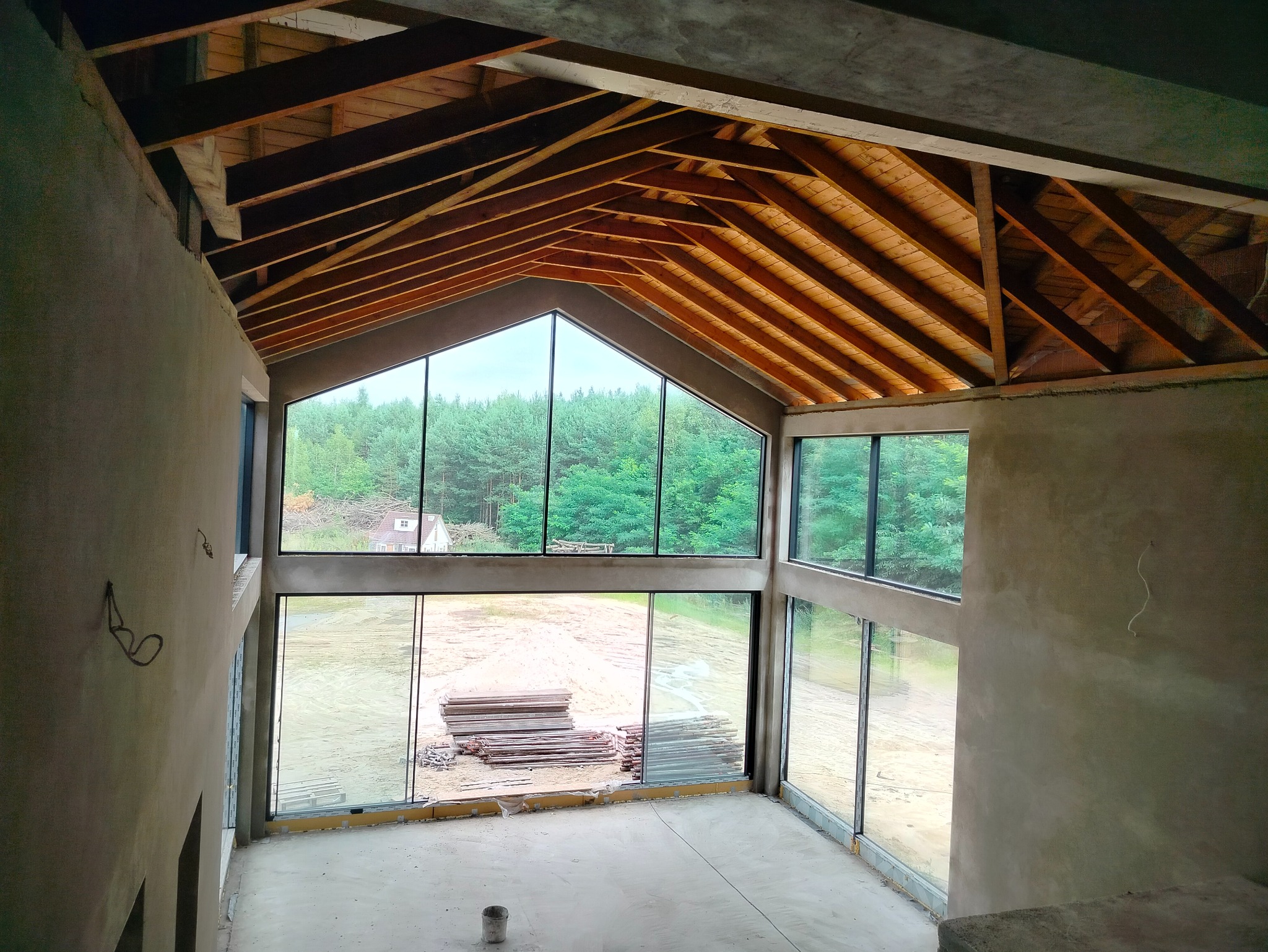 Wnętrze domu w stanie surowym otwartym: surowe ściany, drewniana konstrukcja dachu, duże okna z widokiem na las. Na podłodze widoczne materiały budowlane.