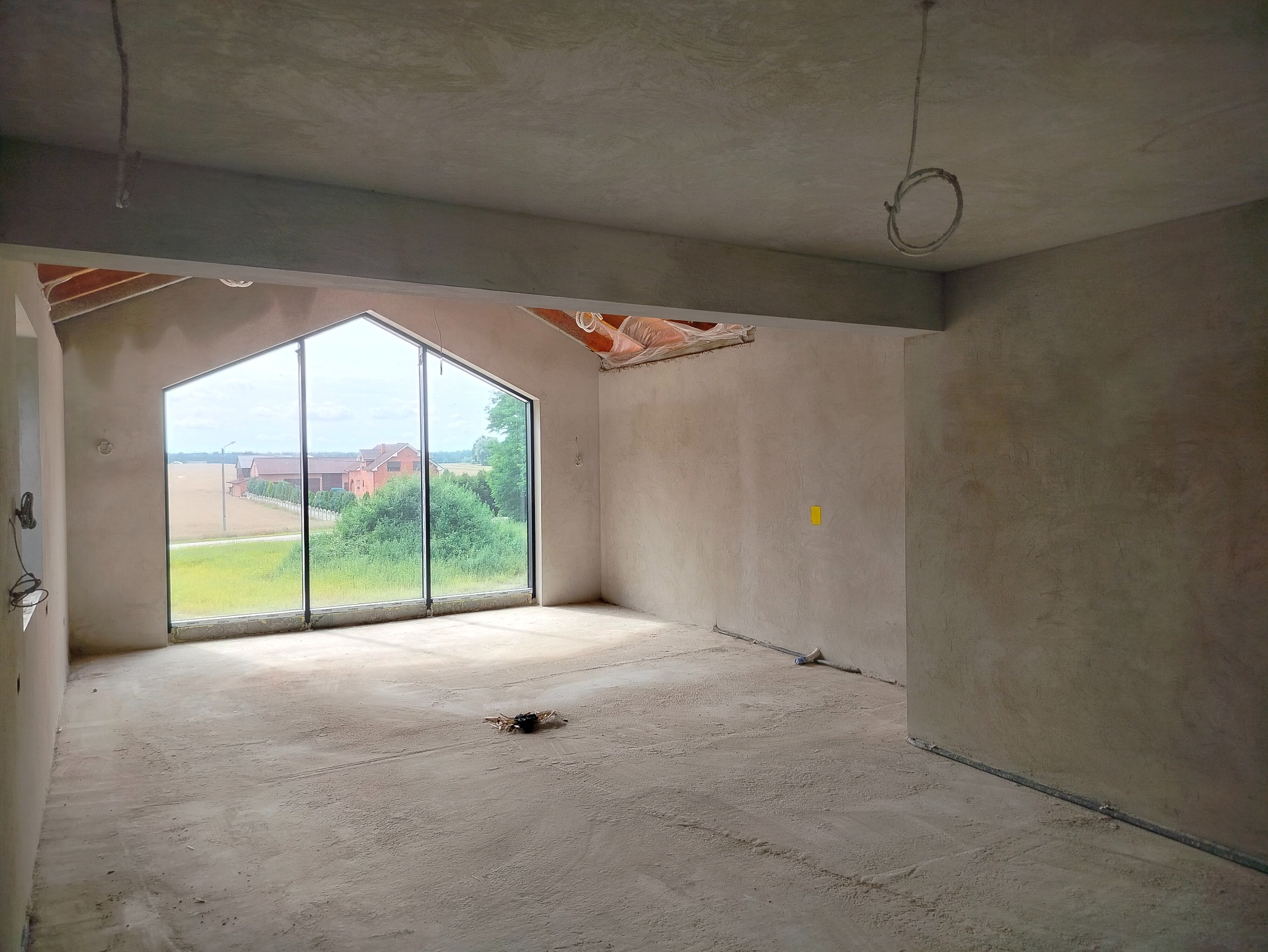 Surowe wnętrze z dużym oknem w kształcie trapezu, ściany pokryte świeżym tynkiem, widoczne przewody elektryczne. Podłoga betonowa, w tle krajobraz wiejski.