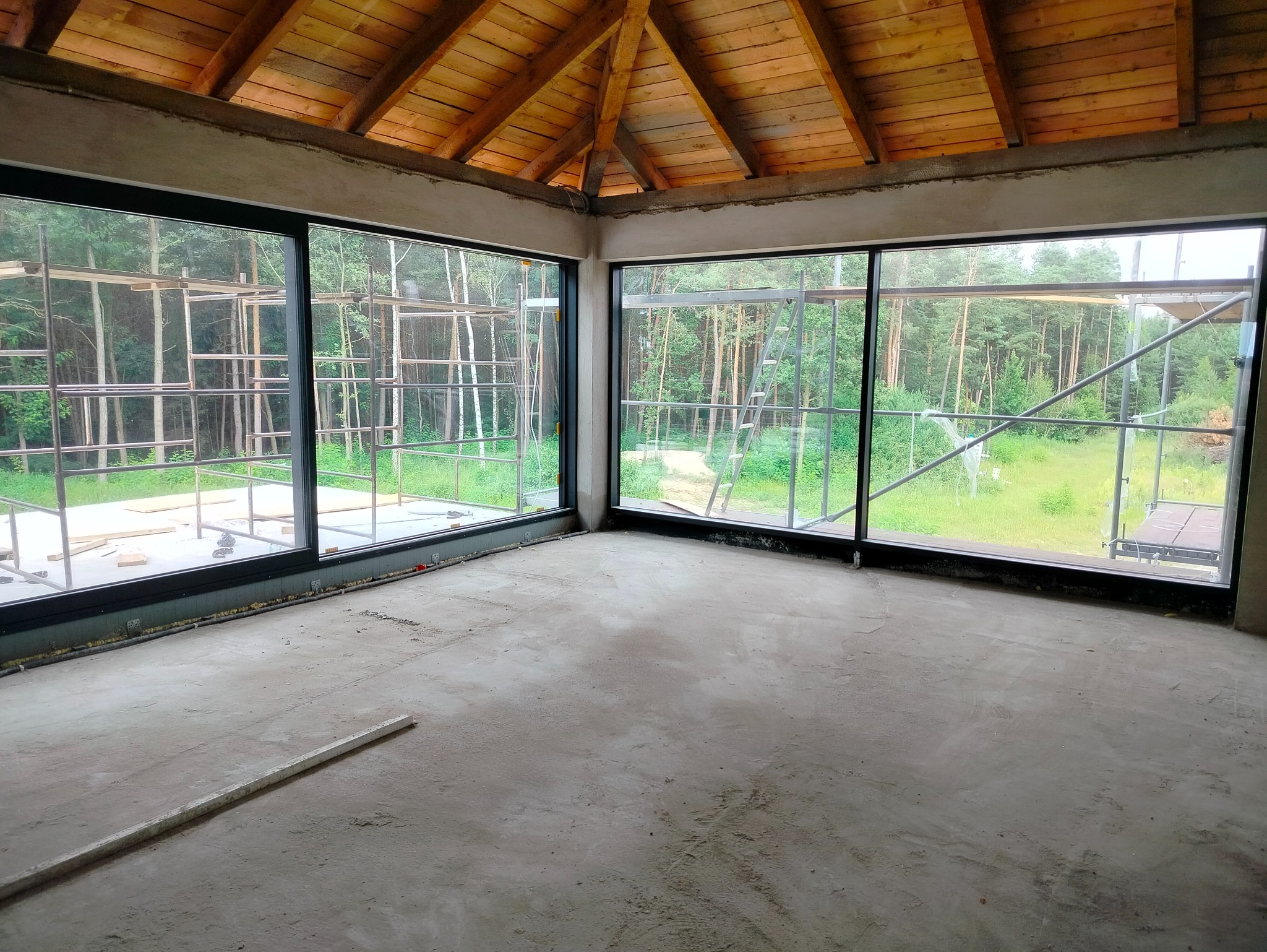 Surowy stan wnętrza z dużymi oknami i drewnianym stropem. Widoczne rusztowania na zewnątrz, betonowa podłoga i ślady po pracach wykończeniowych na ścianach.
