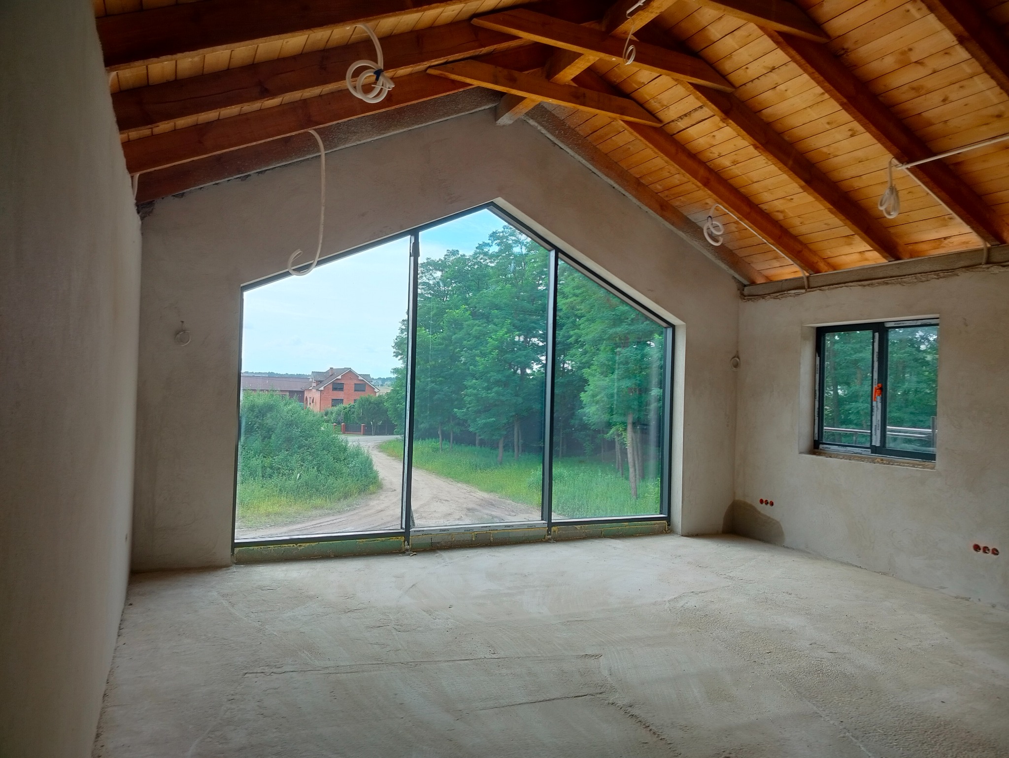 Surowe wnętrze poddasza z drewnianą więźbą dachową, dużym oknem trójkątnym i świeżo położonymi tynkami. Widoczne wystające przewody elektryczne.