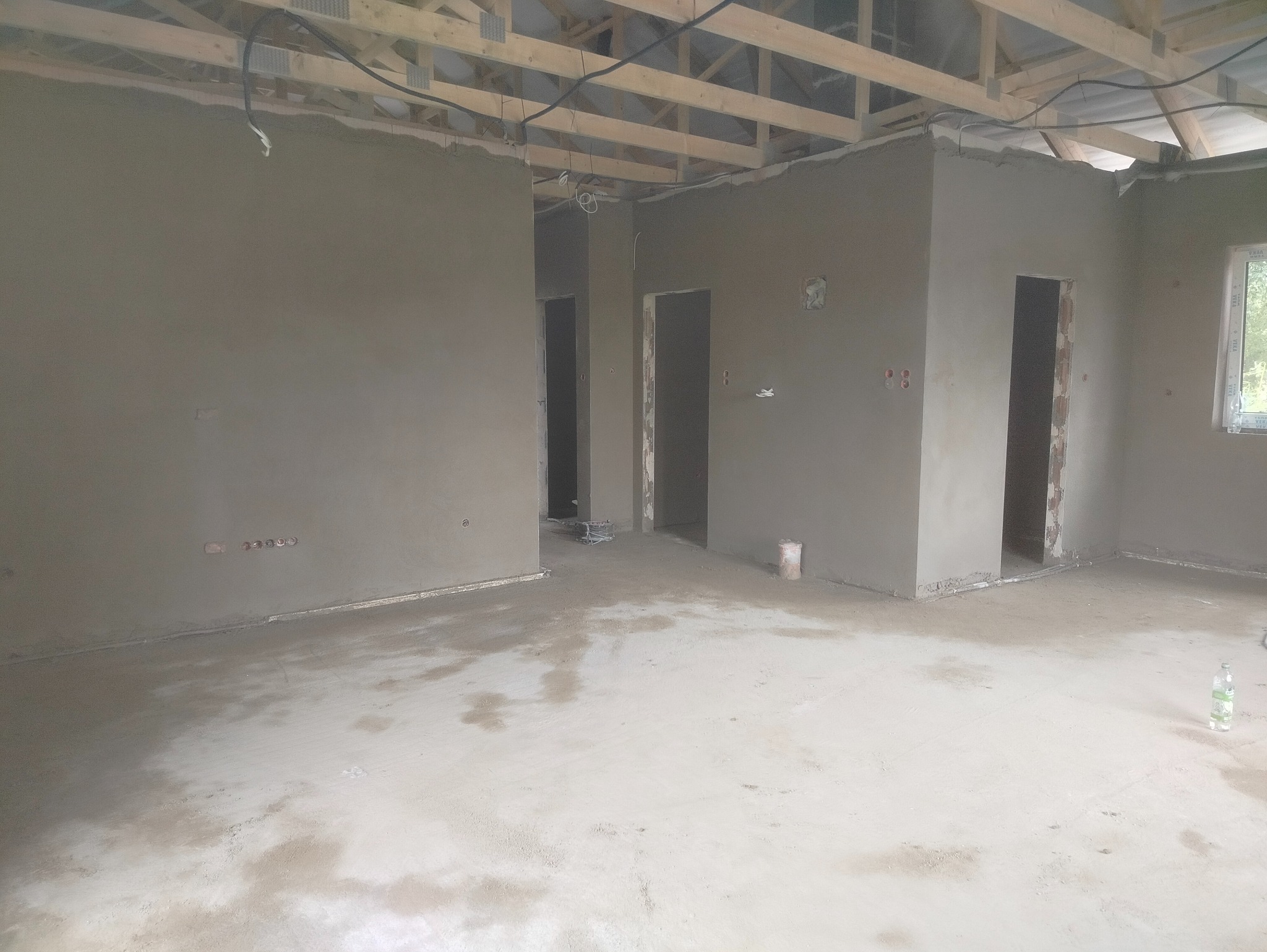 Wnętrze w trakcie prac tynkarskich: świeżo otynkowane ściany, widoczne instalacje elektryczne, surowy strop drewniany, przygotowane otwory drzwiowe. Jasne, naturalne światło.