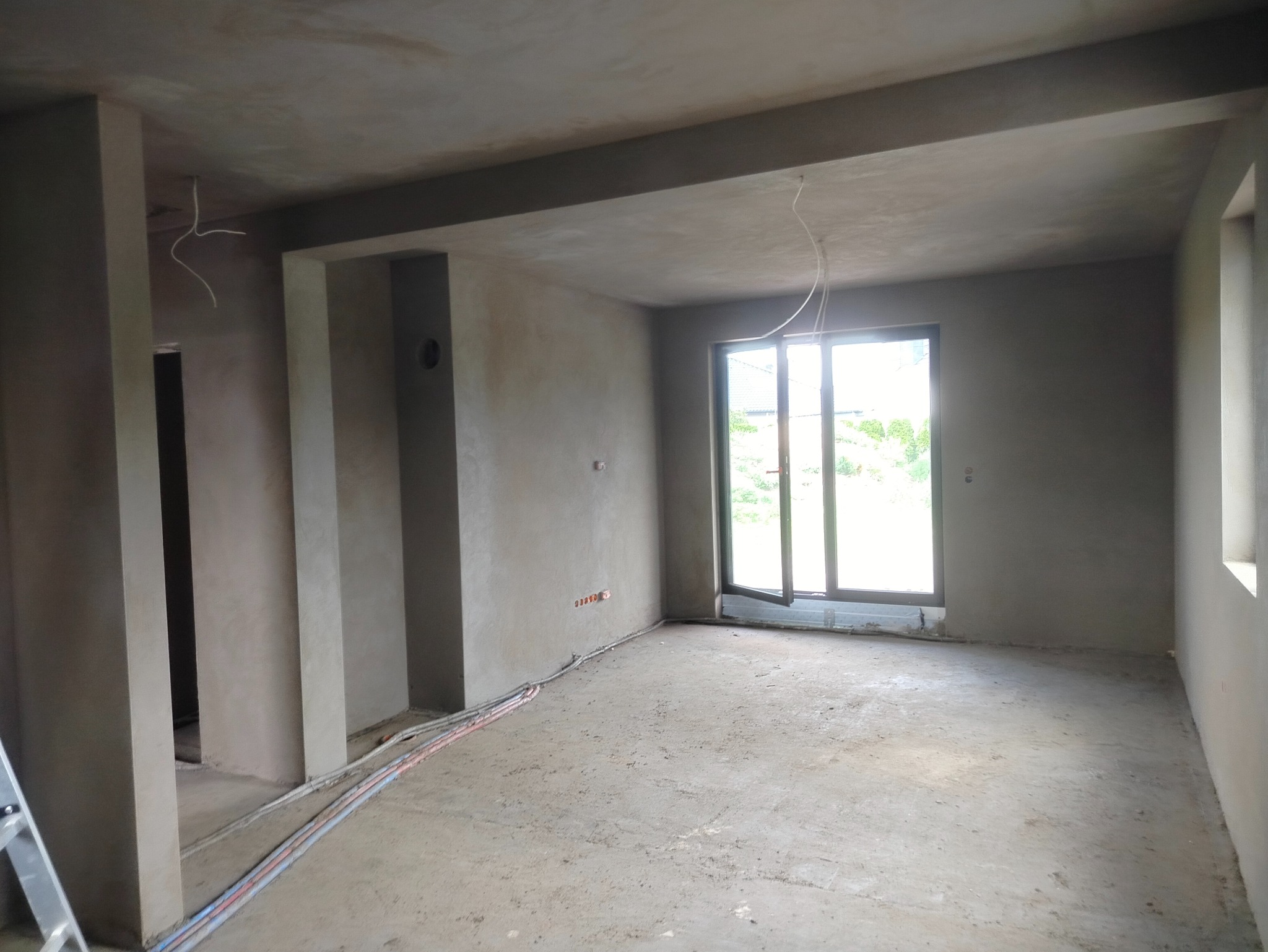 Wnętrze po tynkowaniu maszynowym w Koninie. Świeżo nałożone tynki na ścianach, widoczne kable elektryczne. Duże okno z widokiem na zieleń.