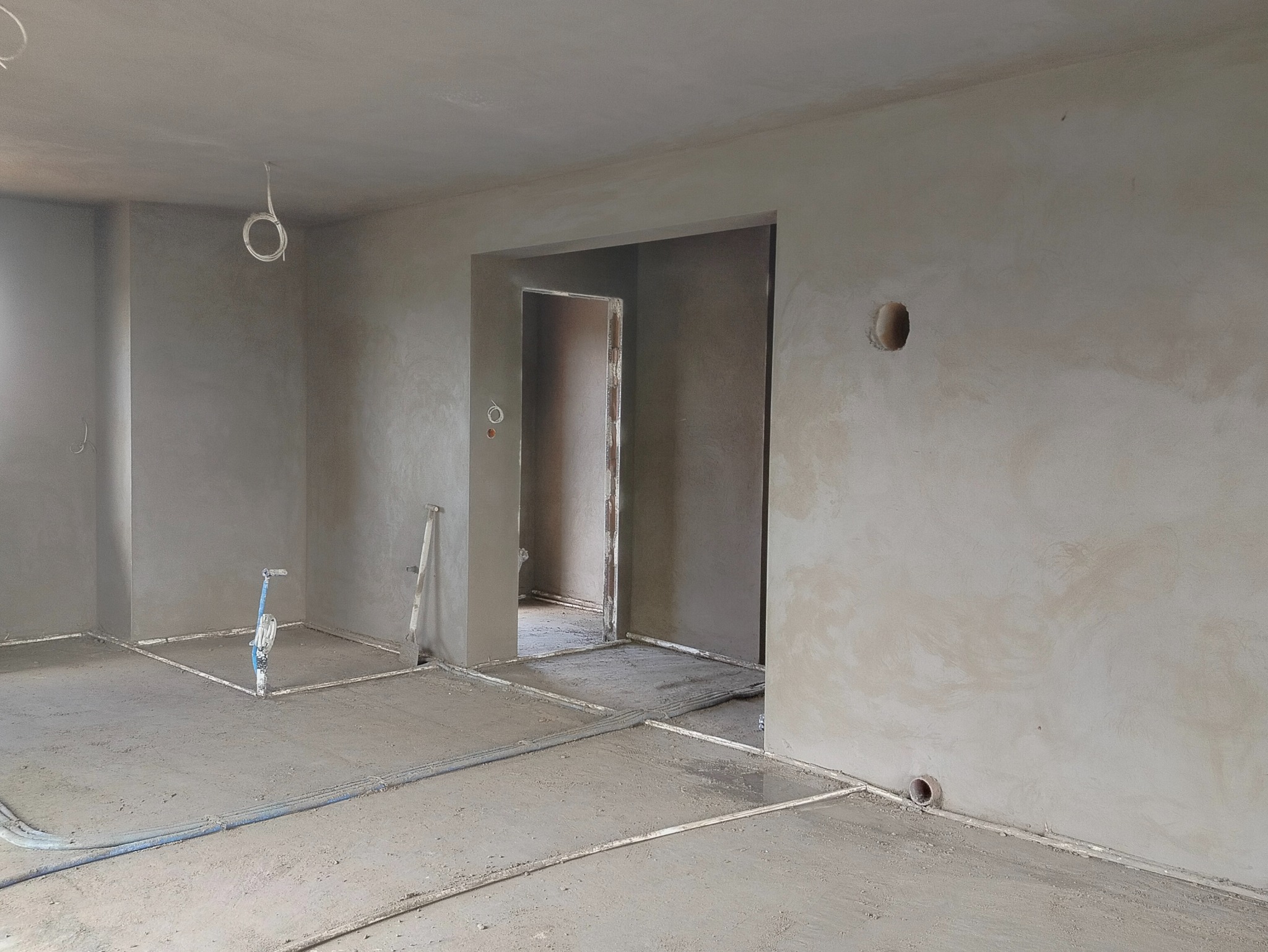 Świeżo otynkowane wnętrze z widocznymi instalacjami podłogowymi i otworem w ścianie, gotowe do dalszych prac wykończeniowych. Szpachla na ścianach i suficie.