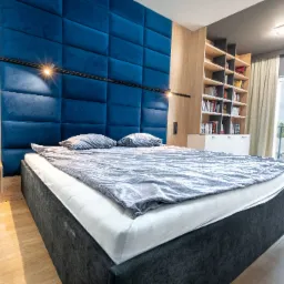 Nowoczesna sypialnia z łóżkiem z ciemną ramą i szarą pościelą, niebieski panel tapicerowany za wezgłowiem, drewniana szafka nocna i regał z książkami przy oknie.