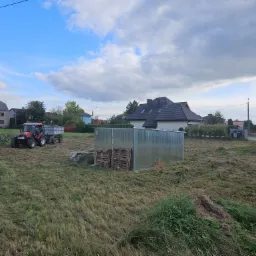 Czerwony traktor z przyczepą na polu po koszeniu trawy, widoczny blaszany garaż i domy w tle, częściowo skoszona trawa.