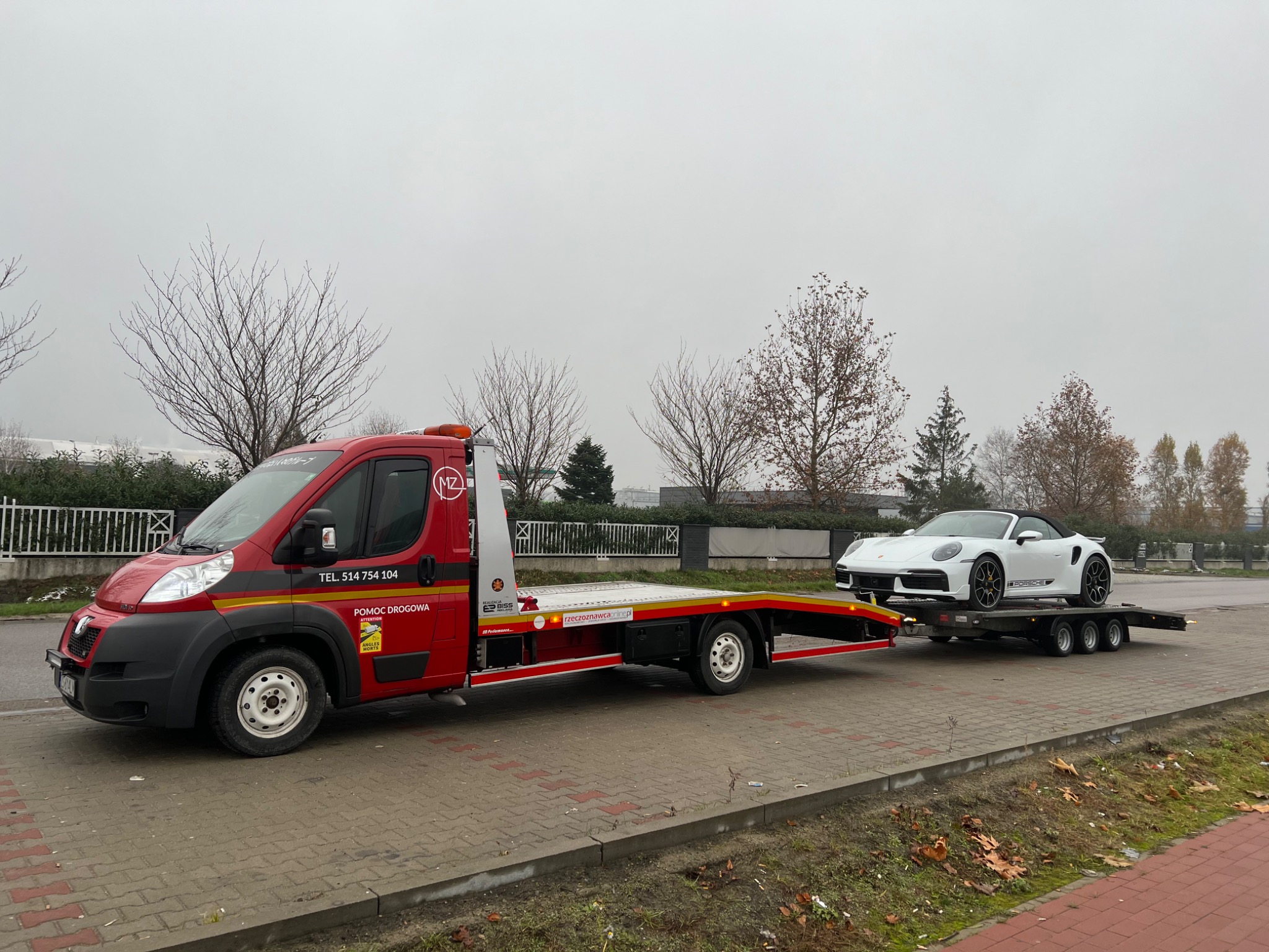 Czerwona laweta z białym Porsche 911 Turbo S Cabriolet na platformie, widok z boku na tle pochmurnego nieba i drzew bez liści.