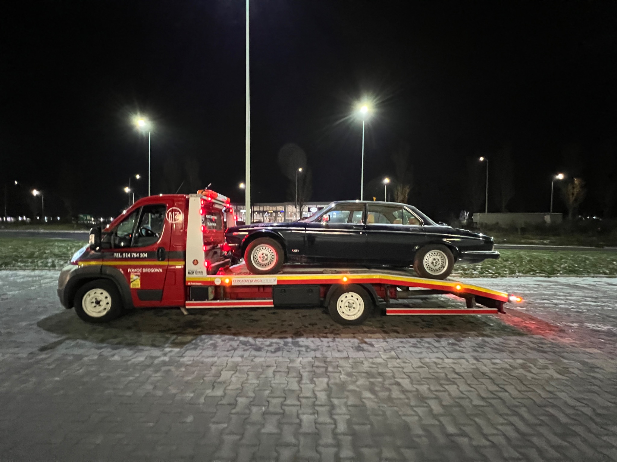 Czerwona laweta z czarnym, klasycznym Jaguarem na platformie, oświetlona światłami ulicznymi w nocy. Widoczny napis 'Pomoc Drogowa' na pojeździe.