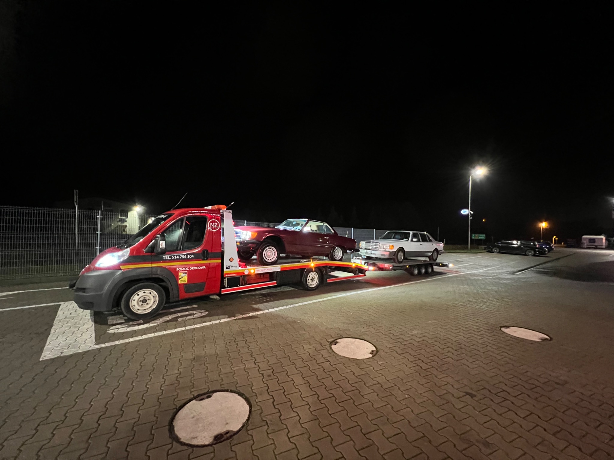 Czerwona laweta z dwoma klasycznymi samochodami Mercedes na parkingu nocą, oświetlona latarnią uliczną.