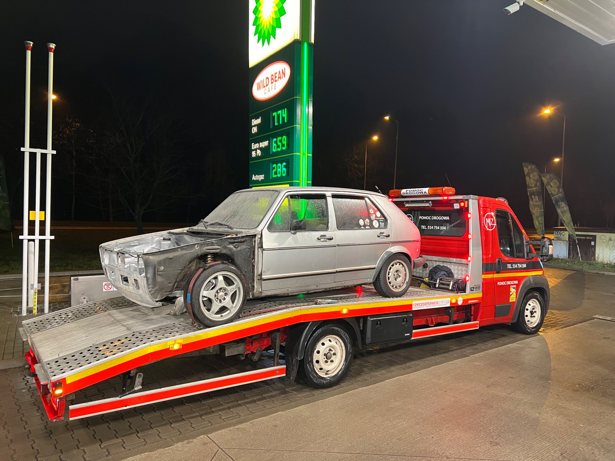 Uszkodzony, częściowo rozebrany srebrny Volkswagen Golf na czerwonej lawecie pomocy drogowej na stacji benzynowej BP nocą, widoczny cennik paliw.