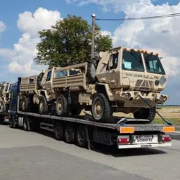 Ciężarówka transportowa przewozi na naczepie kilka wojskowych pojazdów w kolorze khaki, zabezpieczonych pasami transportowymi. W tle widoczne budynki i błękitne niebo z chmurami.