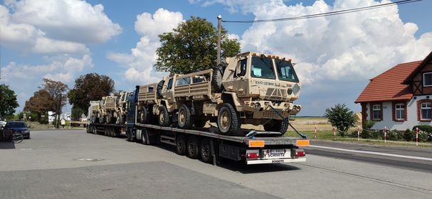 Ciężarówka transportowa przewozi na naczepie kilka wojskowych pojazdów w kolorze khaki, zabezpieczonych pasami transportowymi. W tle widoczne budynki i błękitne niebo z chmurami.