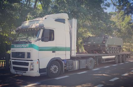 Biała ciężarówka Volvo z zielonym pasem 'TRANSPORT' przewożąca wojskowy transporter opancerzony na platformie, widok z boku na tle drzew.