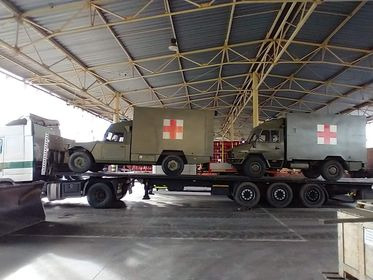 Dwa wojskowe ambulanse z czerwonymi krzyżami, załadowane na niskopodwoziową naczepę ciężarówki, widok z boku w hali magazynowej.