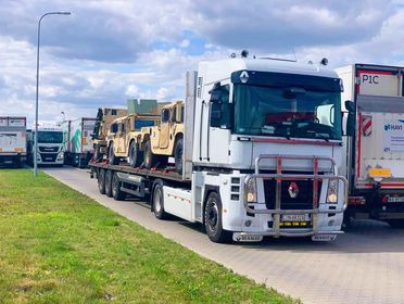 Ciężarówka z naczepą przewożąca kilka wojskowych pojazdów Humvee w kolorze piaskowym na tle zieleni i błękitnego nieba z chmurami.