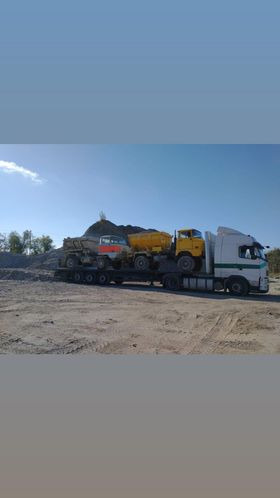 Ciężarówka transportująca dwa wywrotki na niskopodwoziowej naczepie, widok z boku na placu budowy, z hałdą piasku w tle.