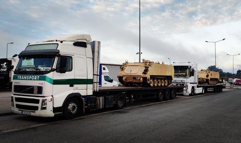 Dwa białe ciągniki siodłowe z naczepami typu laweta przewożące pojazdy wojskowe w kolorze piaskowym na parkingu z oświetleniem ulicznym.