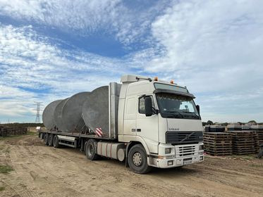 Ciężarówka Volvo FH przewożąca na platformie trzy duże, okrągłe zbiorniki ze stali nierdzewnej, zabezpieczone pasami, widoczne znaki ostrzegawcze na ładunku, tło stanowią pola i słupy wysokiego...