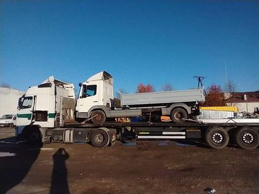 Ciężarówka transportowa przewożąca mniejszy pojazd ciężarowy na platformie, widoczne niebieskie niebo i drzewa w tle, zarys sylwetki osoby robiącej zdjęcie w dolnym rogu.