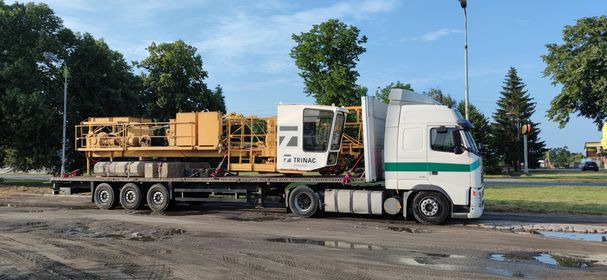 Biała ciężarówka z zielonym pasem transportuje żółty, częściowo zdemontowany dźwig budowlany marki Trinac na niskopodwoziowej platformie. Widoczne betonowe przeciwwagi. Tło stanowią drzewa...