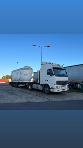 Biała ciężarówka Volvo z naczepą przewozi duży, biały zbiornik z logotypem na tle błękitnego nieba i szarej nawierzchni. Widoczne lampy uliczne i fragmenty innych pojazdów.