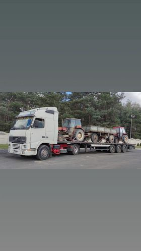 Biała ciężarówka z naczepą przewożąca dwa czerwone traktory rolnicze na tle drzew i betonowego płotu.