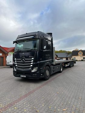 Czarny ciągnik siodłowy Mercedes-Benz Actros z naczepą niskopodwoziową na brukowanym placu, widok z przodu lekko z boku, pochmurne niebo w tle.