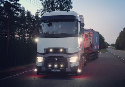 Ciężarówka Renault z naczepą transportująca kontener z pojazdem w tle, sfotografowana z przodu na drodze asfaltowej o zmierzchu.