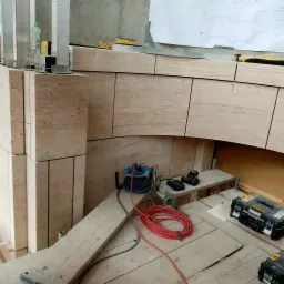 Układanie beżowych płytek trawertynowych w łuk nad otworem, widoczne narzędzia glazurnicze i kable na drewnianym rusztowaniu.