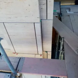 Montaż jasnych, trawertynowych płyt elewacyjnych na rusztowaniu, widoczne elementy mocujące i fragment konstrukcji rusztowania.