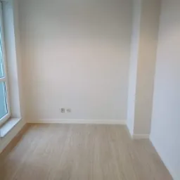 Pusty pokój po remoncie: świeżo pomalowane białe ściany, jasna drewniana podłoga, żółta lampa sufitowa i gniazdka elektryczne przy listwie przypodłogowej.