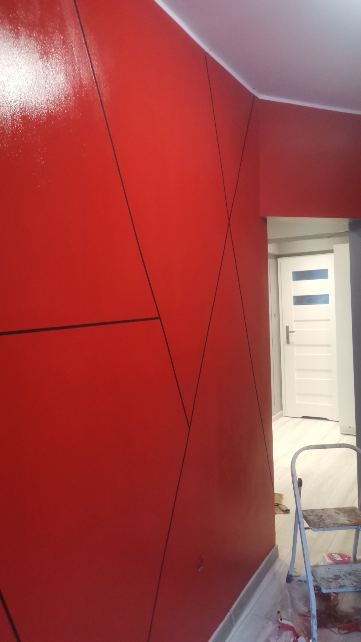 Czerwona ściana z geometrycznym wzorem z czarnych linii, widoczny fragment drabiny malarskiej i białe drzwi w tle.