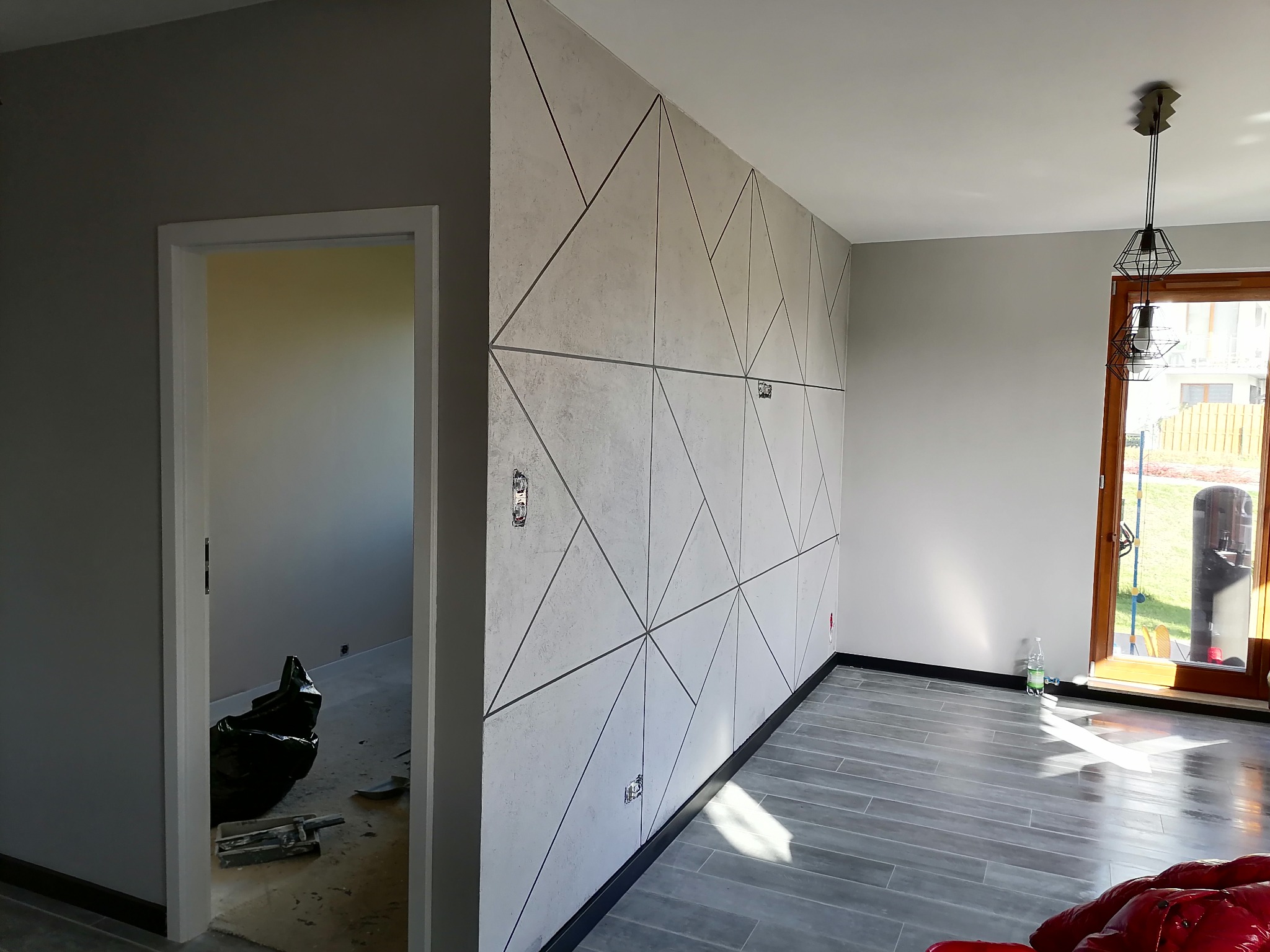 Nowoczesne wykończenie wnętrza z geometryczną ścianą z gładzi, szare ściany i podłoga imitująca drewno, widoczne gniazdka elektryczne.