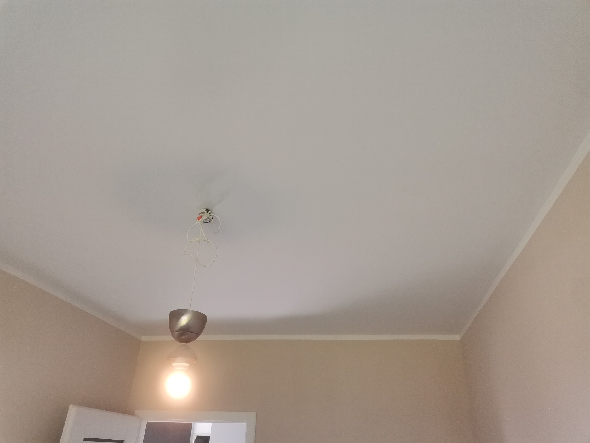 Biały, gładki sufit z widocznym fragmentem instalacji elektrycznej i nowoczesną lampą wiszącą, w tle beżowa ściana i fragment drzwi.
