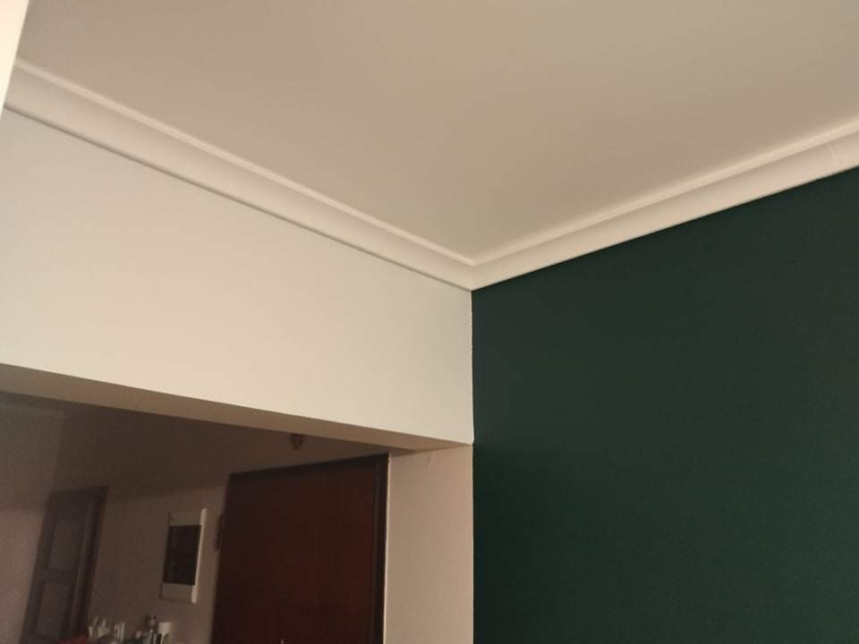 Fragment wnętrza: ciemnozielona ściana pomalowana farbą matową, biały sufit z dekoracyjnym gzymsem, widok na przejście do innego pomieszczenia z ciemnymi drzwiami.