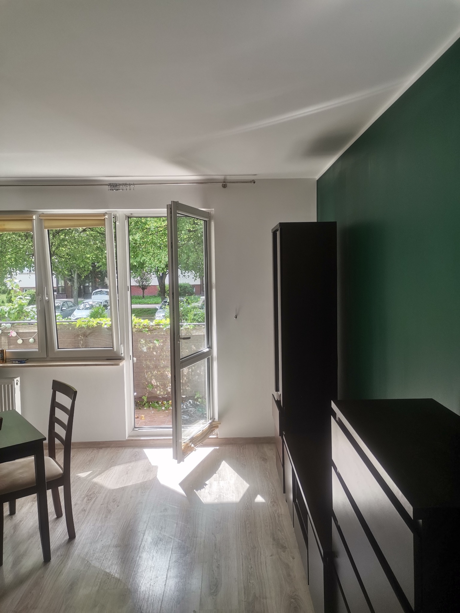 Wnętrze pokoju z otwartym balkonem, świeżo pomalowaną ścianą w kolorze butelkowej zieleni, drewnianą podłogą i meblami w ciemnym kolorze, oświetlone popołudniowym słońcem wpadającym przez okno.