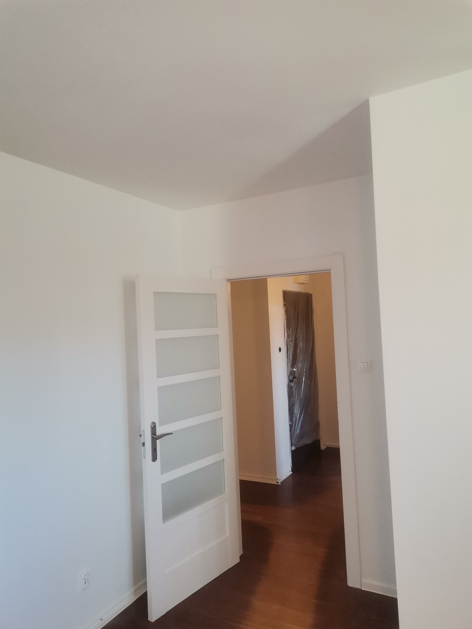 Wnętrze mieszkania po odświeżeniu ścian, widoczna otwarta biała, przeszklona drzwi wewnętrzne oraz podłoga z ciemnego drewna, w tle pomieszczenie zabezpieczone folią malarską.