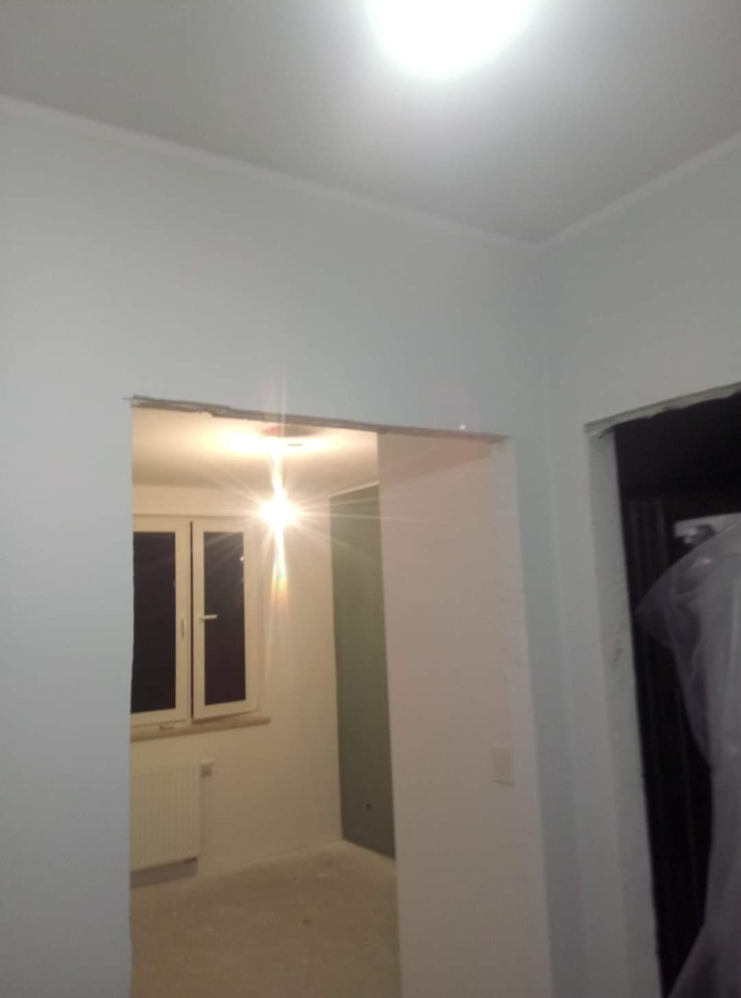 Świeżo pomalowane na biało wnętrze pokoju z widokiem na okno, widoczne ślady po taśmie malarskiej przy futrynie drzwi i listwie sufitowej.