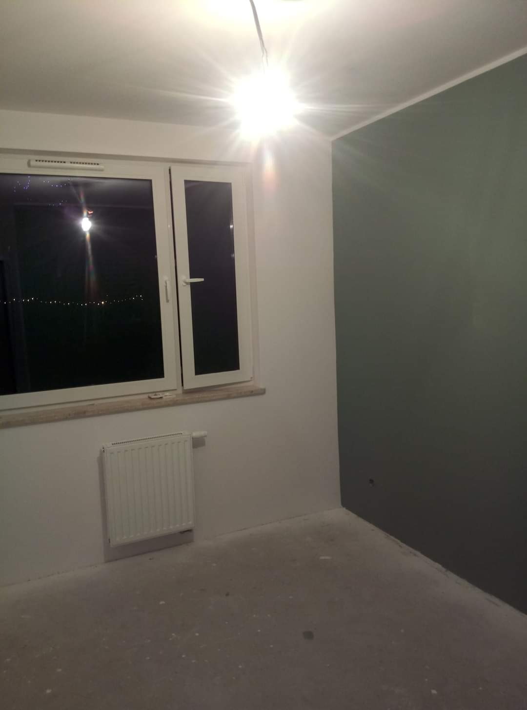 Pomalowane na biało ściany i sufit w pustym pokoju z oknem i grzejnikiem, jedna ściana w kolorze ciemnej zieleni, widoczny kabel od lampy sufitowej.