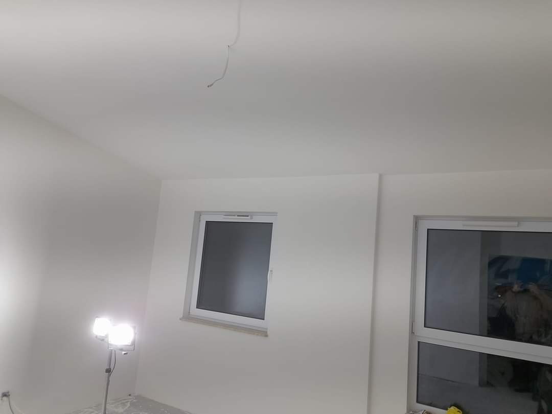 Świeżo pomalowane na biało wnętrze pokoju z dwoma oknami, widoczny przewód zwisający z sufitu i lampa halogenowa na podłodze.