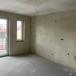 Surowe wnętrze w trakcie remontu w Warszawie: ściany po tynkowaniu maszynowym z widocznymi puszkami elektrycznymi, sufit z widocznymi śladami szpachlowania, szara wylewka na podłodze. Widok przez...