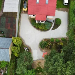 Widok z drona na posesję z jasną kostką brukową tworzącą ścieżki i podjazd, obramowaną ciemniejszymi kostkami i otoczoną zielenią, z widocznym czerwonym dachem domu i zaparkowanymi samochodami.