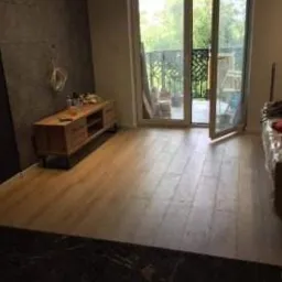 Jasna drewniana podłoga w trakcie układania, widoczne przygotowane panele i narzędzia, ściana z szarą okładziną, otwarte drzwi balkonowe z widokiem na zieleń.