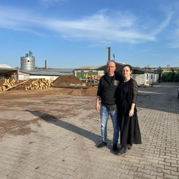 Studio Dekoracji Wnętrz Forrest - Para na tle składu drewna z pociętymi kłodami i budynkami przemysłowymi w tle, w słoneczny dzień. Widoczny stos drewna i przyczepa.