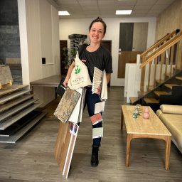 Studio Dekoracji Wnętrz Forrest - Uśmiechnięta kobieta w salonie z próbkami podłóg, płytek i listew przypodłogowych. Widoczne schody i ekspozycja materiałów wykończeniowych.
