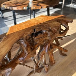 Studio Dekoracji Wnętrz Forrest - Unikatowy blat drewniany z korzenia, o nieregularnym kształcie i bogatej fakturze, oświetlony promieniami słońca. W tle okrągły stół z blatem z żywicy i kwiatami.