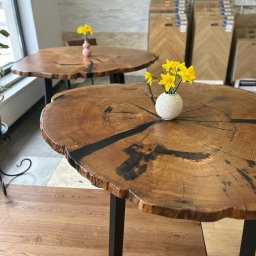 Studio Dekoracji Wnętrz Forrest - Dwa okrągłe stoliki kawowe z blatem z litego drewna z żywicą epoksydową, na czarnych, metalowych nogach. Na blacie wazonik z żółtymi kwiatami. W tle ekspozycja paneli.